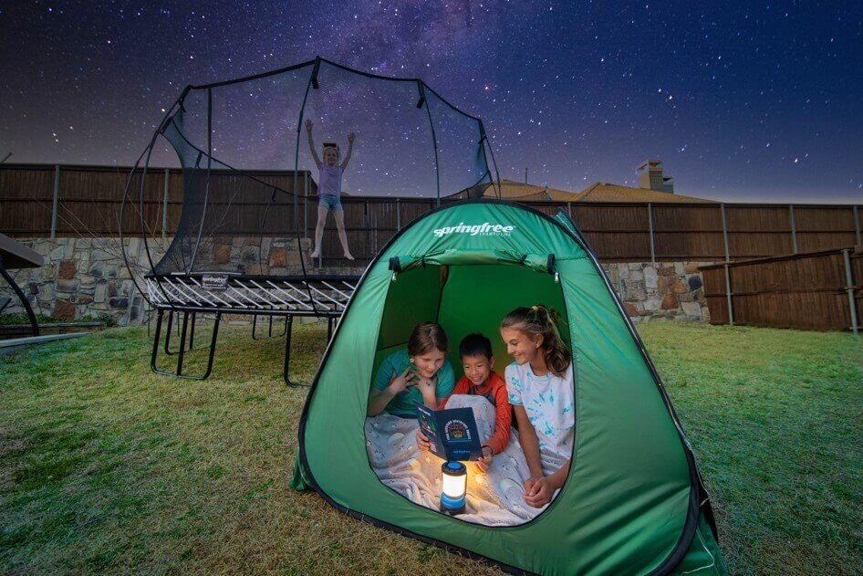 Three kids using the Springfree Backyard Camping Kit while another kid jumps on a trampoline