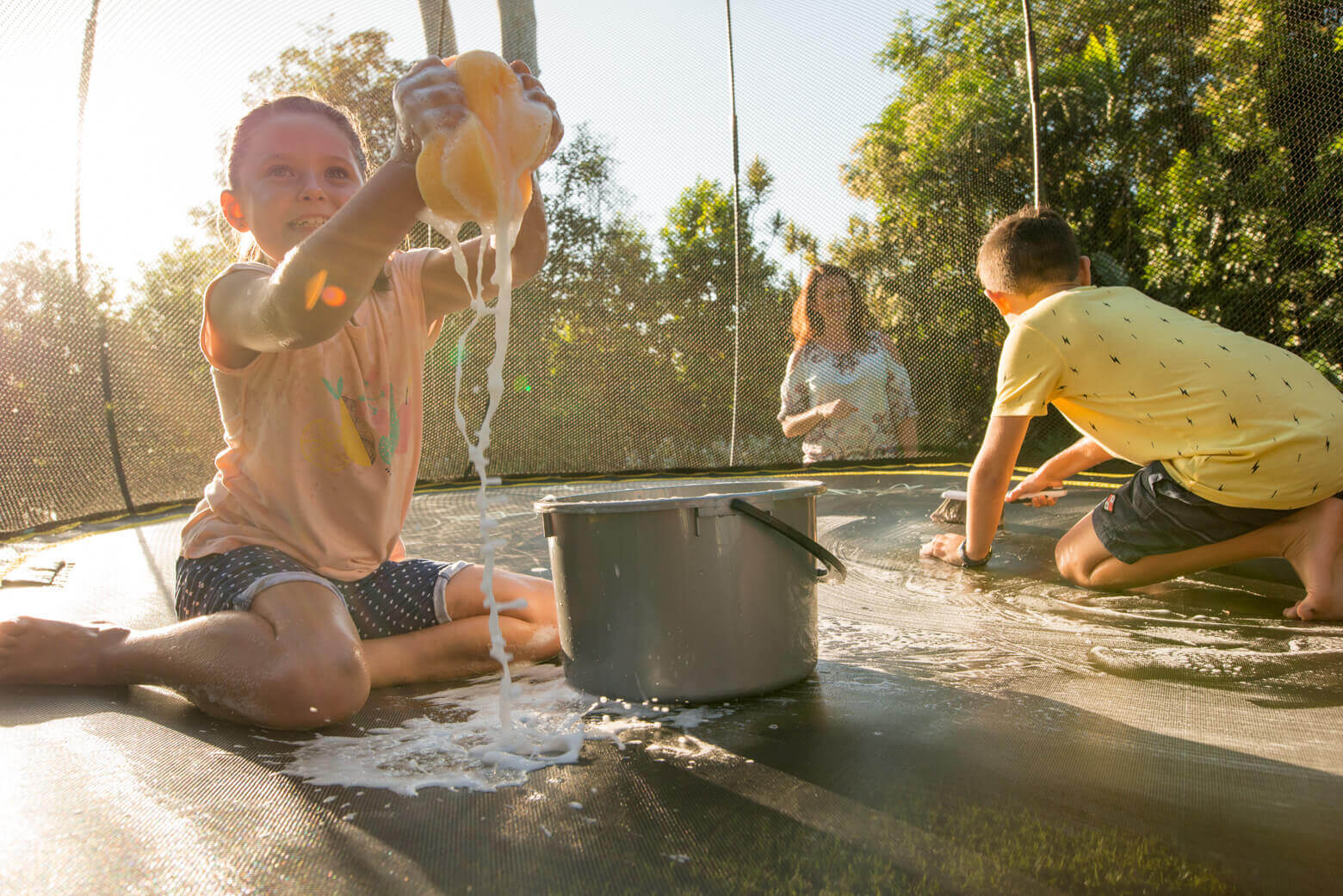 A girl and boy cleaning a trampoline mat with soap and water while their mom watches