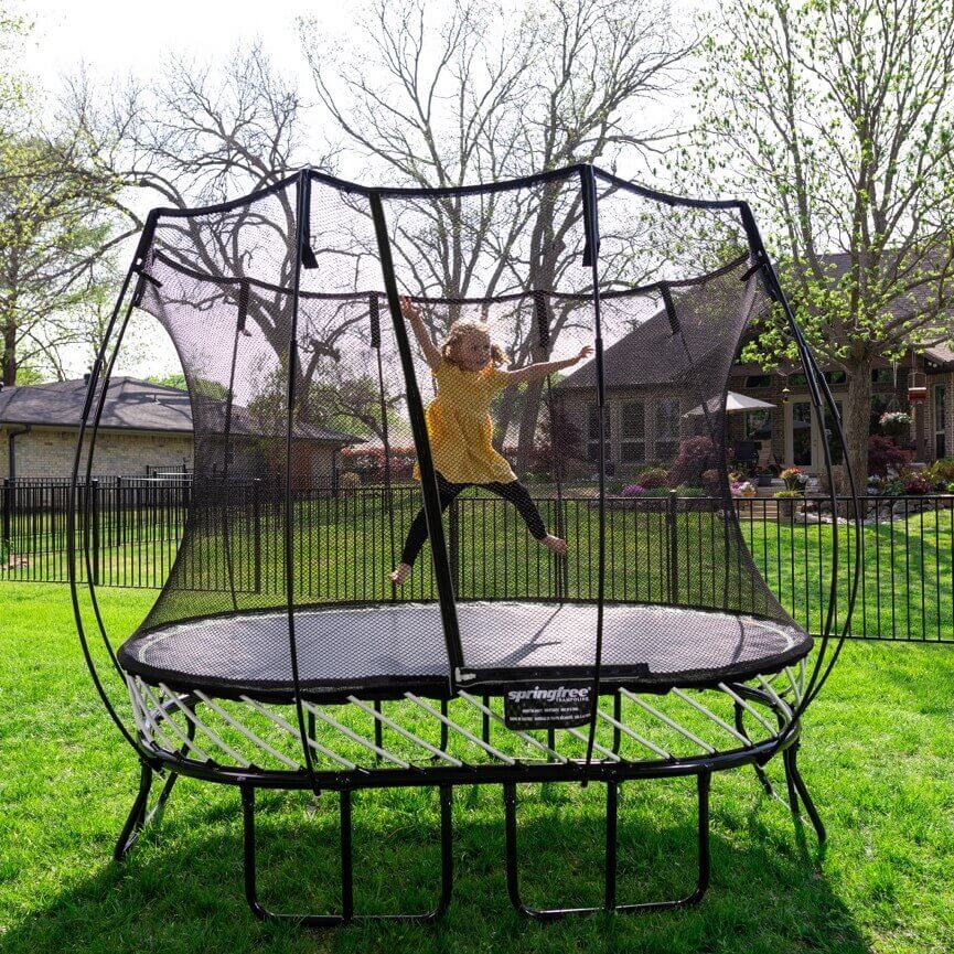 A little girl in a yellow shirt jumping on a Springfree Compact Oval Trampoline.