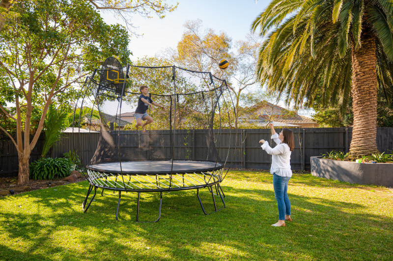 A little boy jumping on a Springfree Medium Round Trampoline catching a ball thrown by his mom.