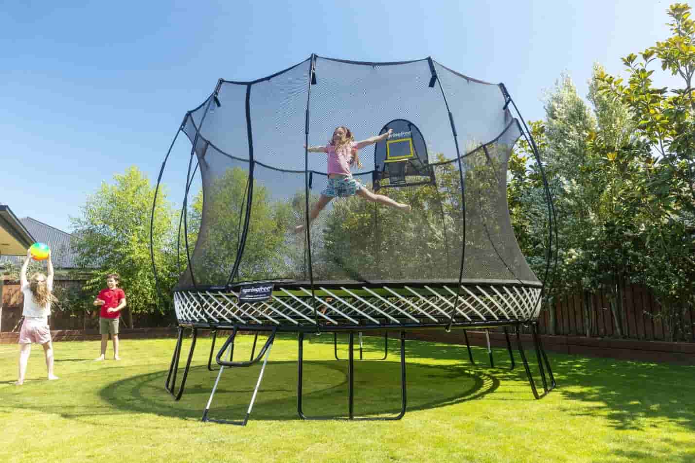 A girl jumping on a trampoline while two other children throw a ball on the side of the trampoline.