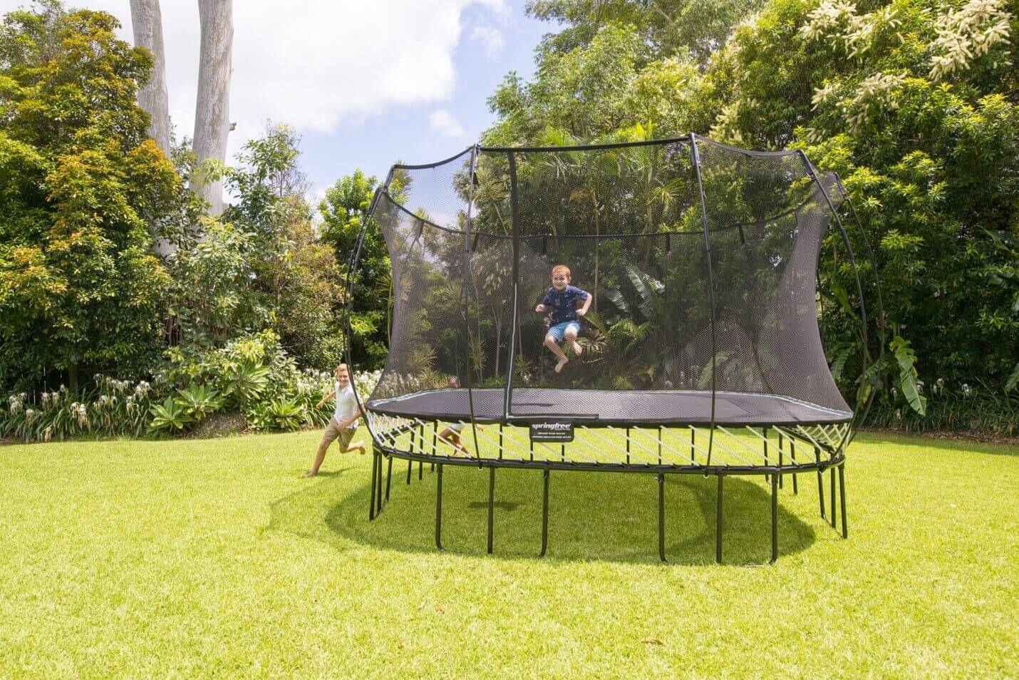 Kid jumping on a Springfree Jumbo Square Trampoline while two other kids chase each other right outside of the trampoline