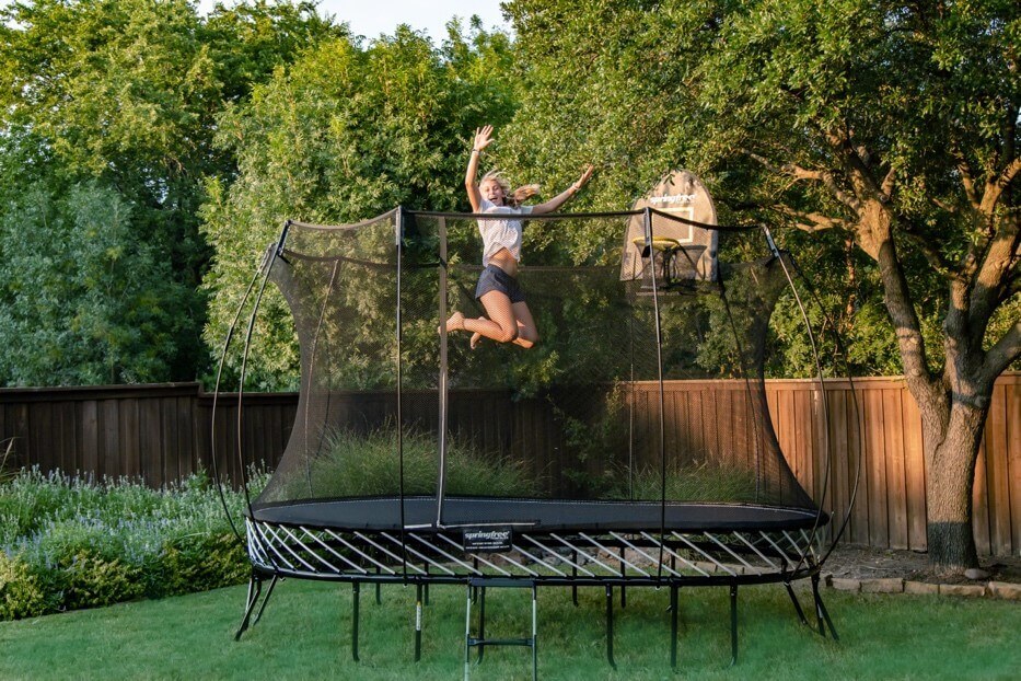 A smiling women jumping on a Springfree Large Oval Trampoline with her hands in the air.