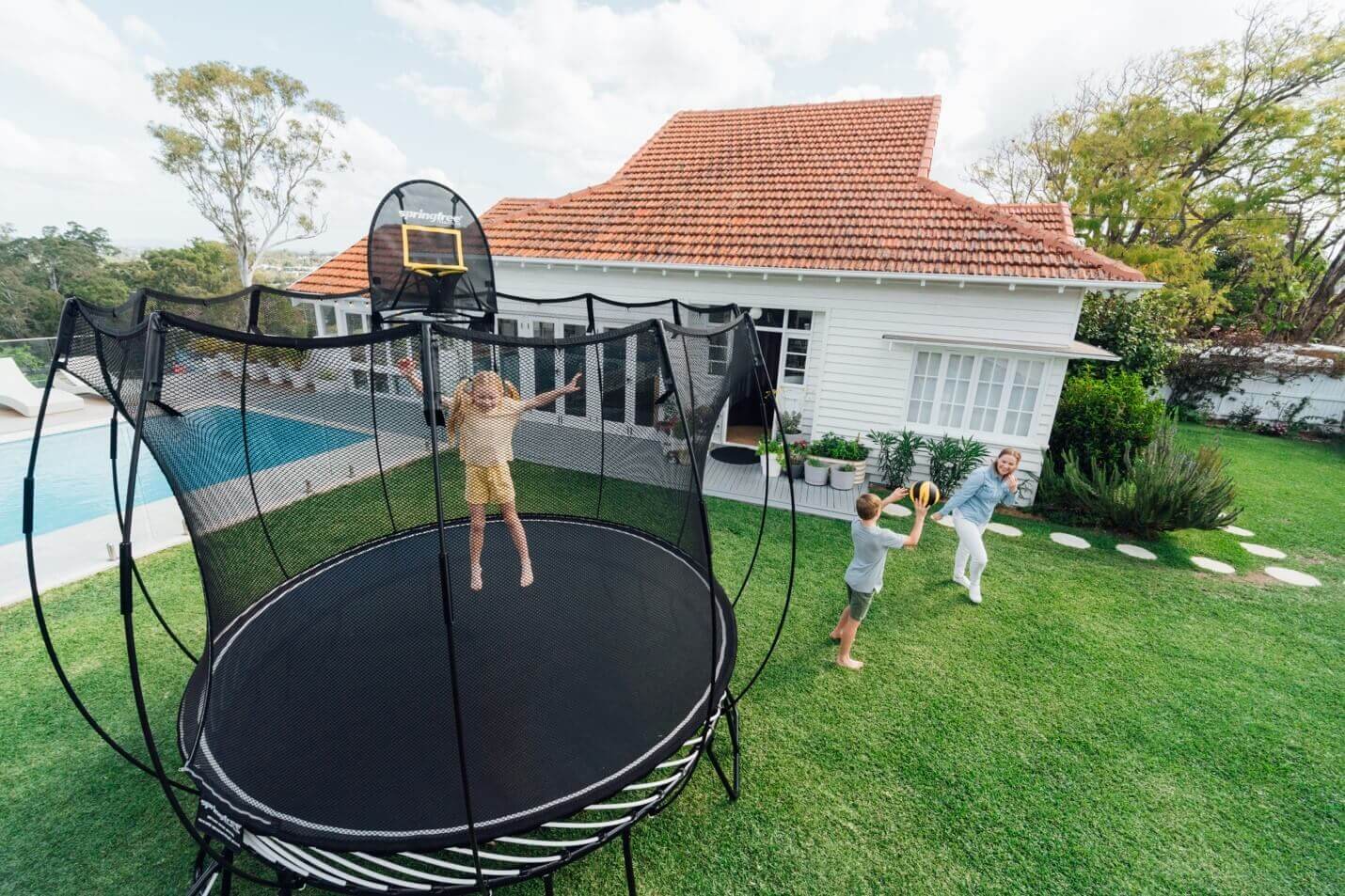 Little girl jumping on a Springfree Trampoline while her brother and mom throw a ball on the outside. 