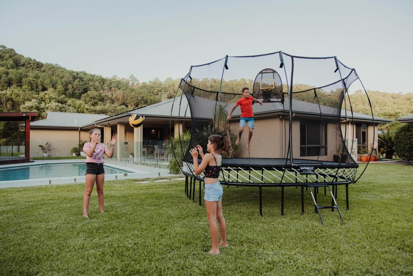 A boy jumping on a Springfree Trampoline while two other kids play ball in the backyard.