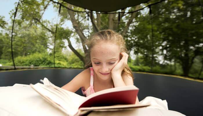 A little girl reading a book on a Springfree Trampoline with a Sunshade attached to the top.