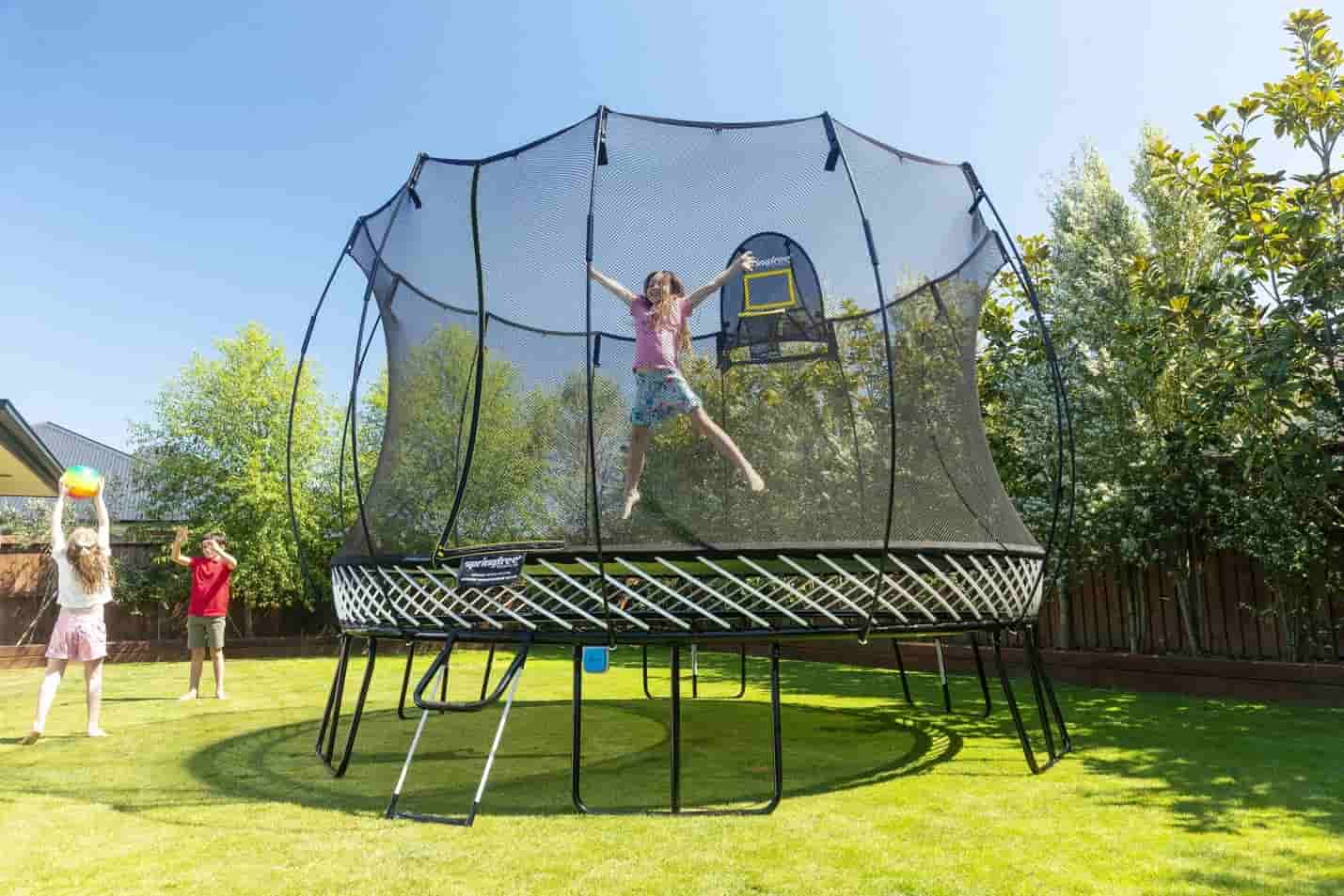 A girl jumping on a Springfree Trampoline while two other kids throw a ball outside of it.