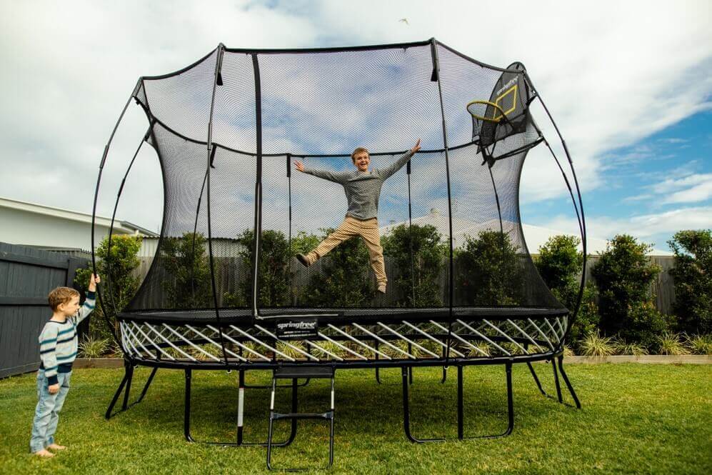 A little kid jumping on a Springfree Large Oval Trampoline while another kid watches from the outside.