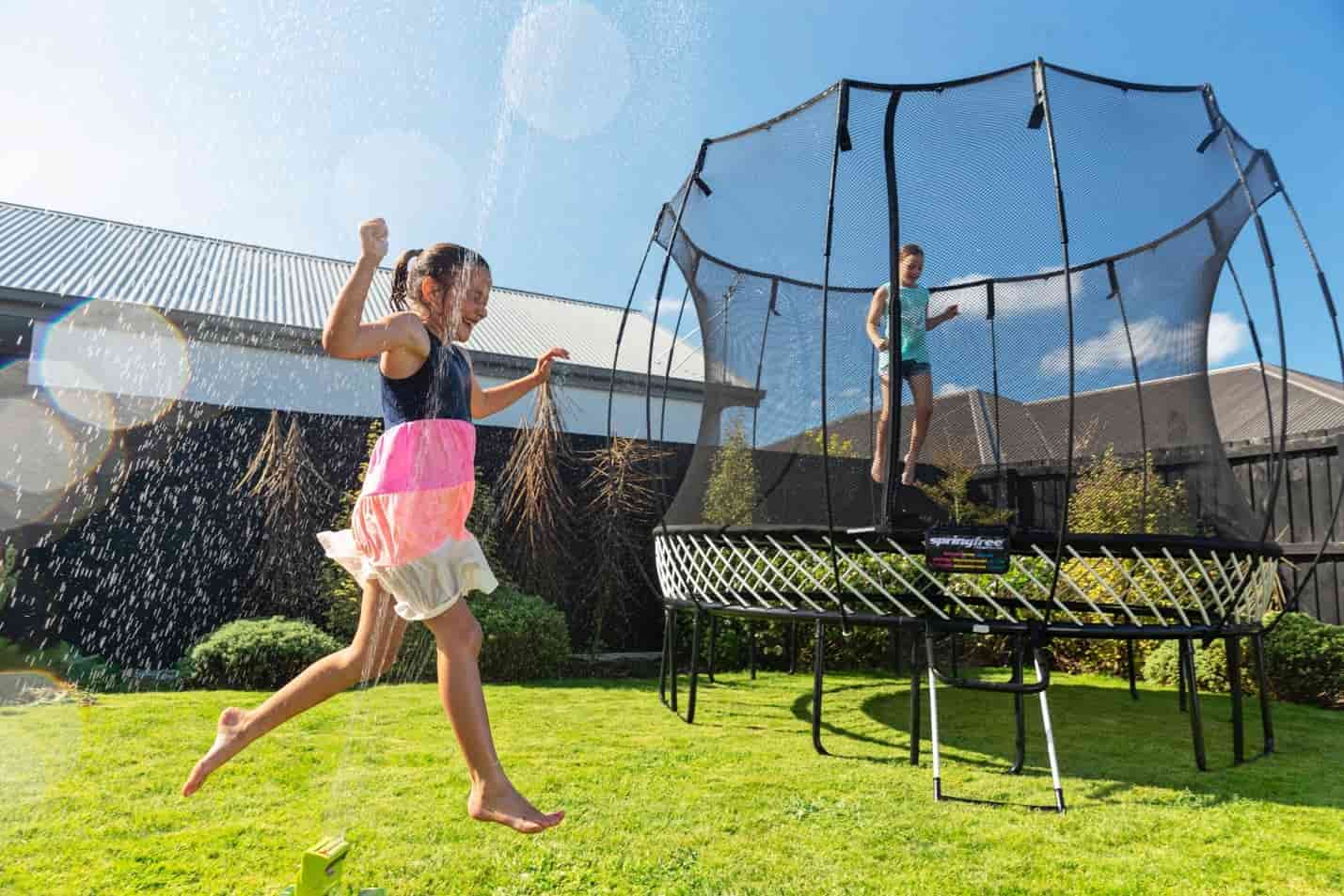 A girl jumping on a Springfree Trampoline while another girl runs through a sprinkler. 