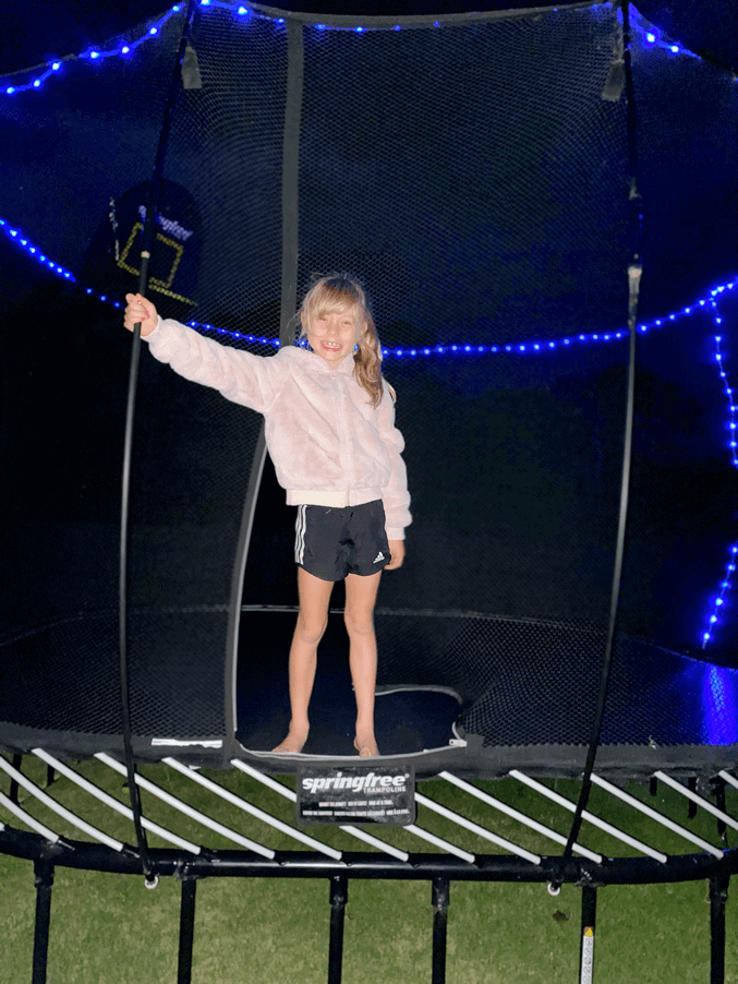 A girl standing on a Springfree Trampoline with string lights attached to it