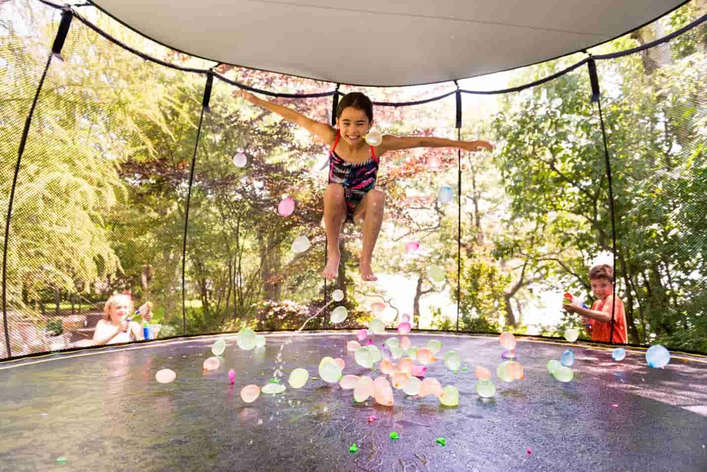 A girl jumping with water balloons on a Springfree Trampoline with a Sunshade attached to the top.