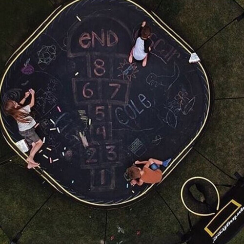 Three kids drawing hopscotch with chalk on a trampoline mat.