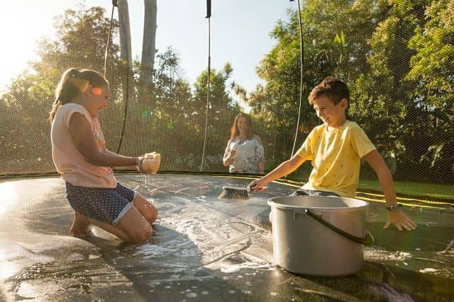 Two kids laughing while cleaning a trampoline with soap and water with their mom watching in the background