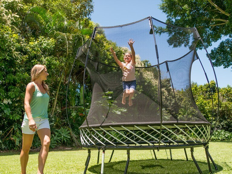 Little girl jumping in mid-air on a trampoline with her mom watching and smiling close by